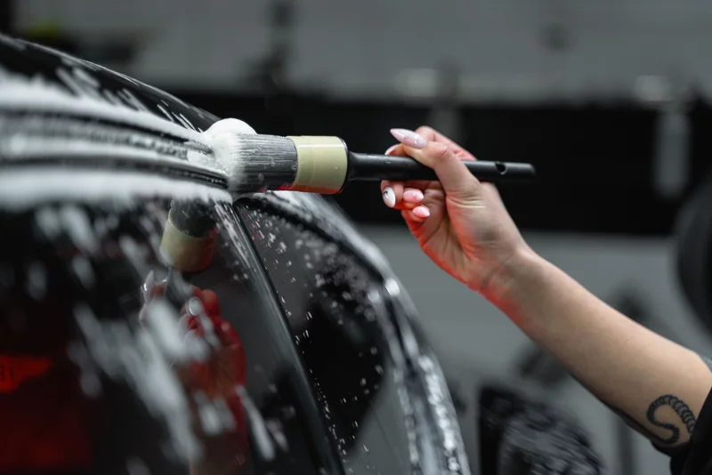A detailing brush cleaning soapy trim beside a car window